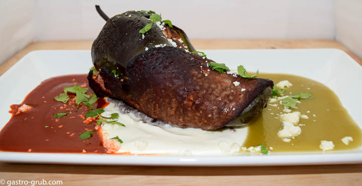 Chile relleno wrapped with flank steak and served with chili sauce, crema, and tomatillo sauce, on a rectangular plate.
