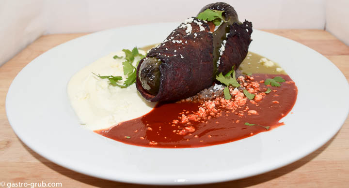 Chile relleno wrapped with flank steak and served with chili sauce, crema, and tomatillo sauce, on a round plate.