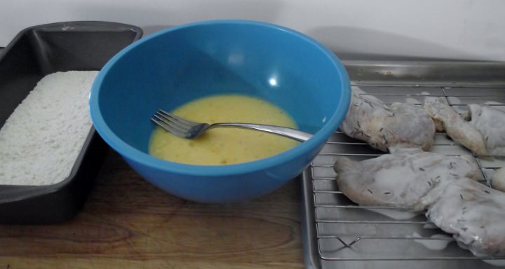 Ingredients for fried chicken: flour in a pan, egg mixture in a bowl, and chicken on a rack.